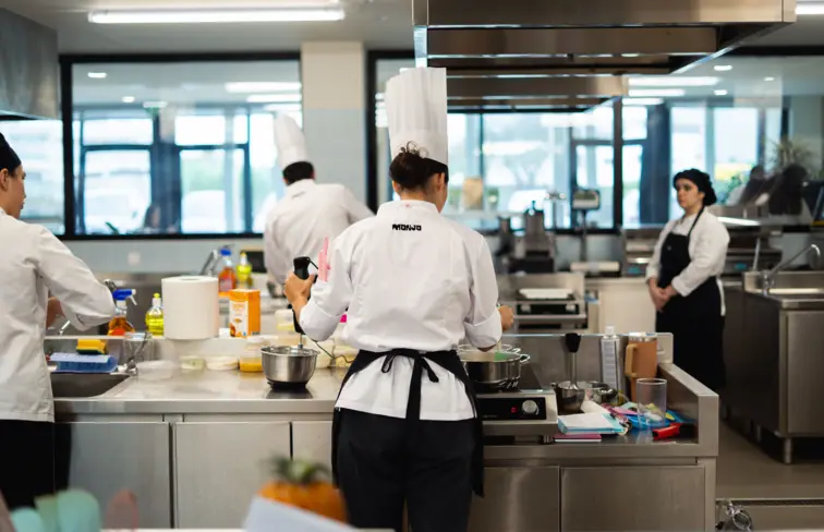 Imagem de uma mulher de costas, numa cozinha, com um chapéu de culinária, junto a uma banca, com vários utensílios de zonha, a cozinhar. Em redor estão outros cozinheiros nas suas tarefas.